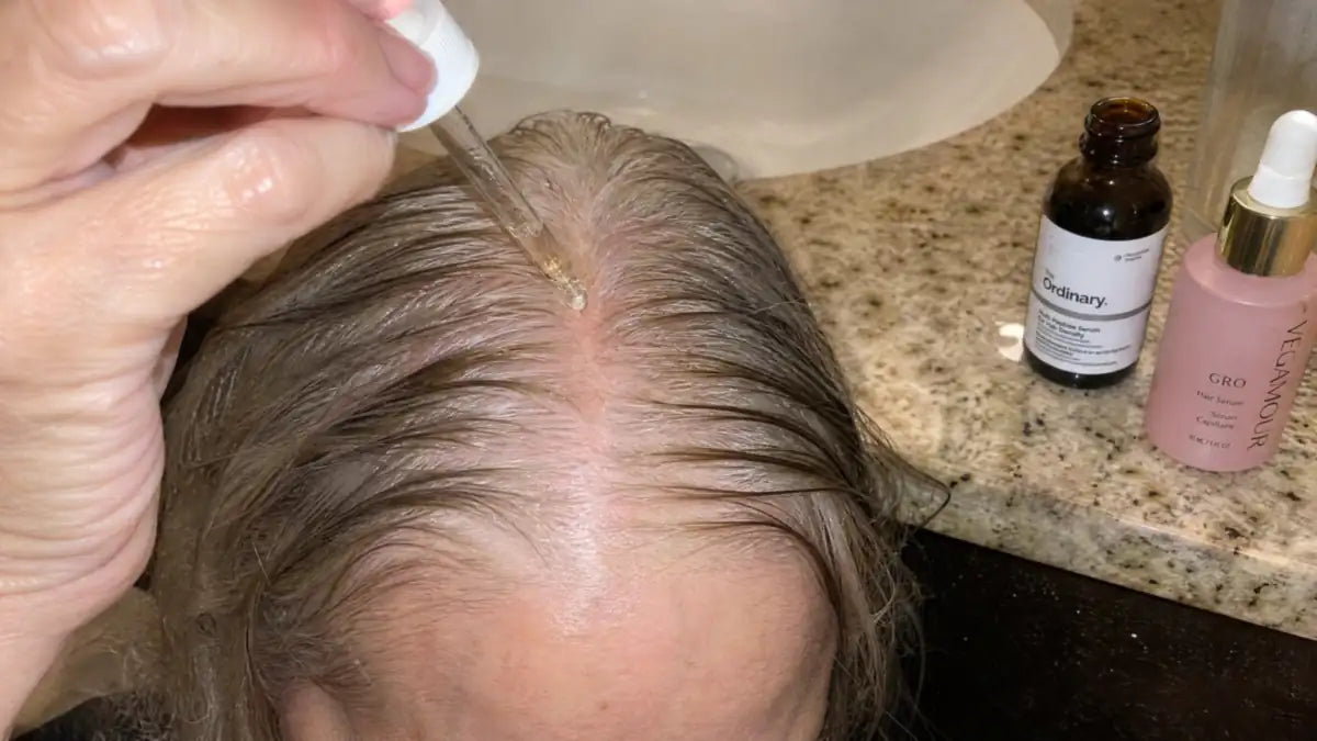 Dropper applying leave-in growth serum to a thinning part, serum bottles on the counter